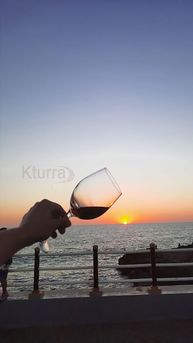 Copa del vino Kturra con la mano extendida hacia la puesta de sol en Poneloya, León.